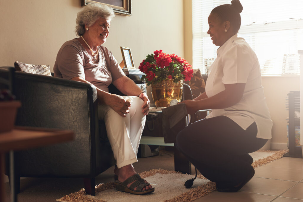 Senior woman sitting on a chair at home with female caregiver holding blood pressure gauge. Female nurse visiting senior patient for checking blood pressure.